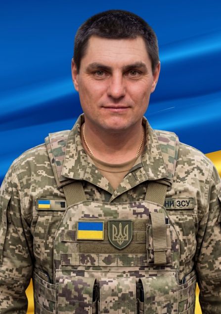 Чоловік у військовій формі Збройних сил України з емблемою та українським прапором на тлі державного прапора України.