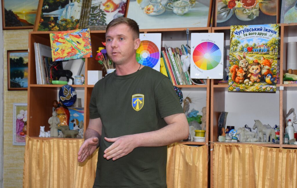 Чоловік у темній зеленій футболці з емблемою на грудях стоїть і жестикулює рукою перед книжковими полицями з різними декоративними предметами та дитячою книжкою із зображенням тварин.