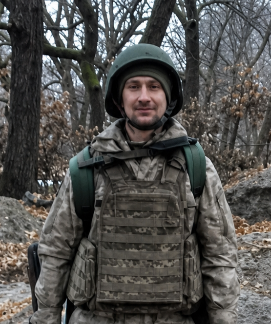 Військовий у камуфляжній формі, шоломі та бронежилеті стоїть серед лісу.