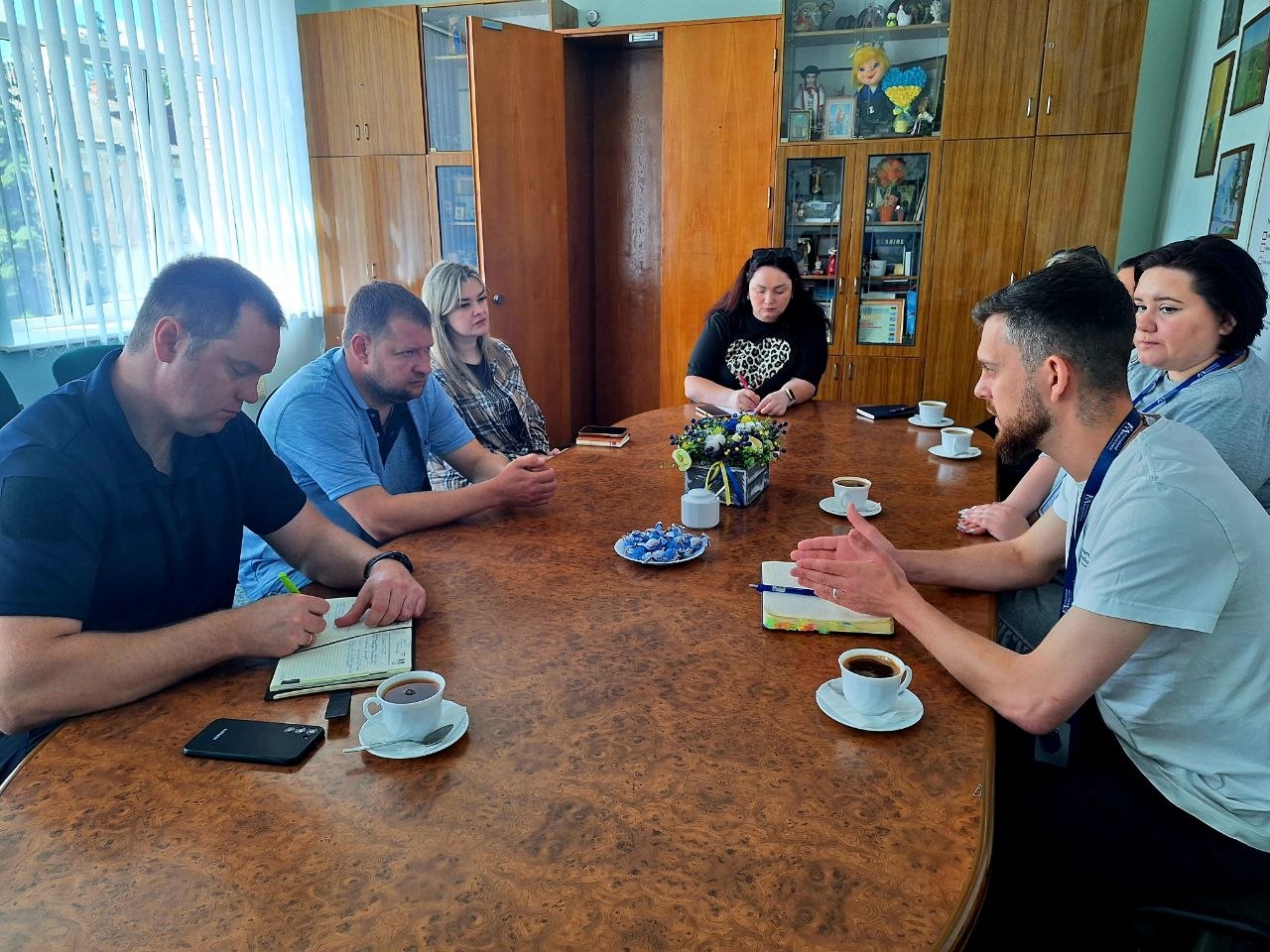 Кілька людей сидять за круглим столом у приміщенні з дерев'яними меблями та обговорюють інформацію, фіксуючи її в нотатках.