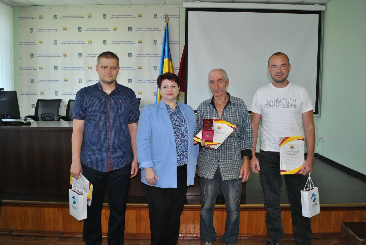 Четверо людей стоять поряд, посміхаючись; двоє з них тримають пакети, а двоє інших - папери та нагороду.