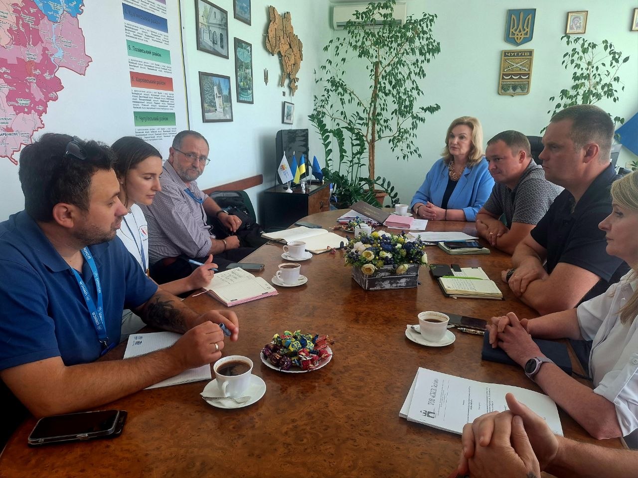 Кілька осіб сидять за столом під час наради чи зустрічі, серед яких чоловік у синій поліоці веде нотатки, а інші уважно слухають.