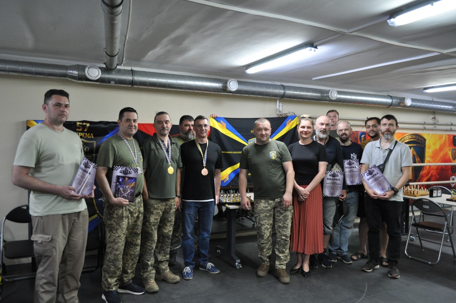 Група людей у військовому одязі та цивільному вбранні зібралась для групового фото, деякі з них тримають нагороди та посвідчення, а на задньому плані видно банери та шахові дошки.