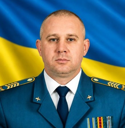 Чоловік у військовій формі із золотими відзнаками на тлі українського прапора.