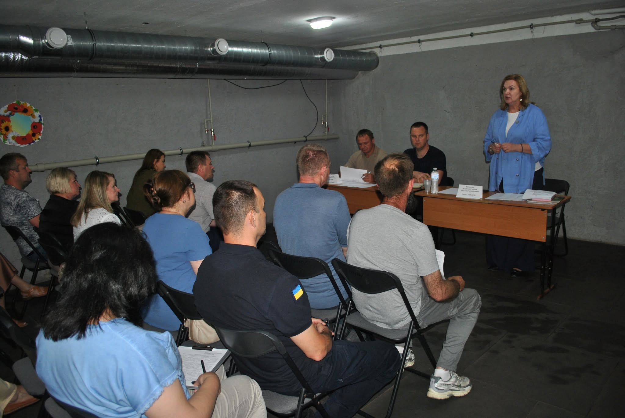 Жінка виступає перед аудиторією, що сидить на стільцях, біля довгого столу з іншими учасниками.