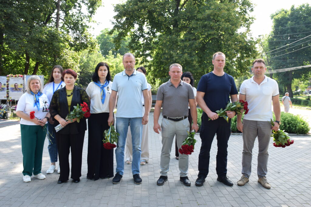 Група людей стоїть у ряд на відкритому повітрі, тримаючи в руках червоні троянди та сині стрічки, ймовірно, під час урочистості вшанування пам'яті.