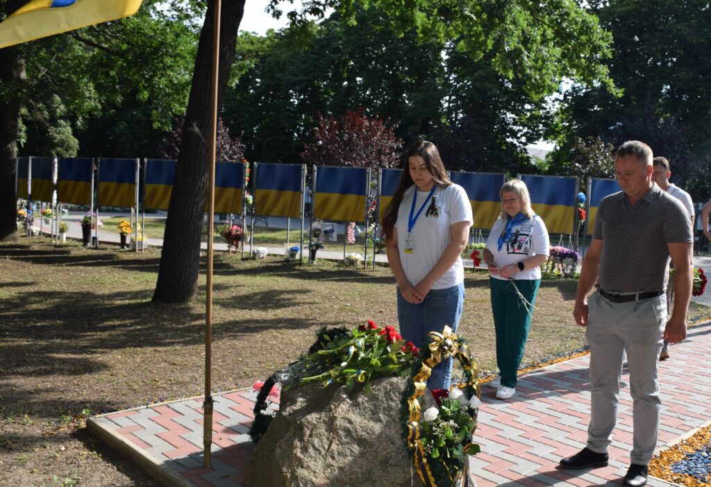Люди стоять біля пам'ятного каменя з квітами на тлі ряду українських прапорів.
