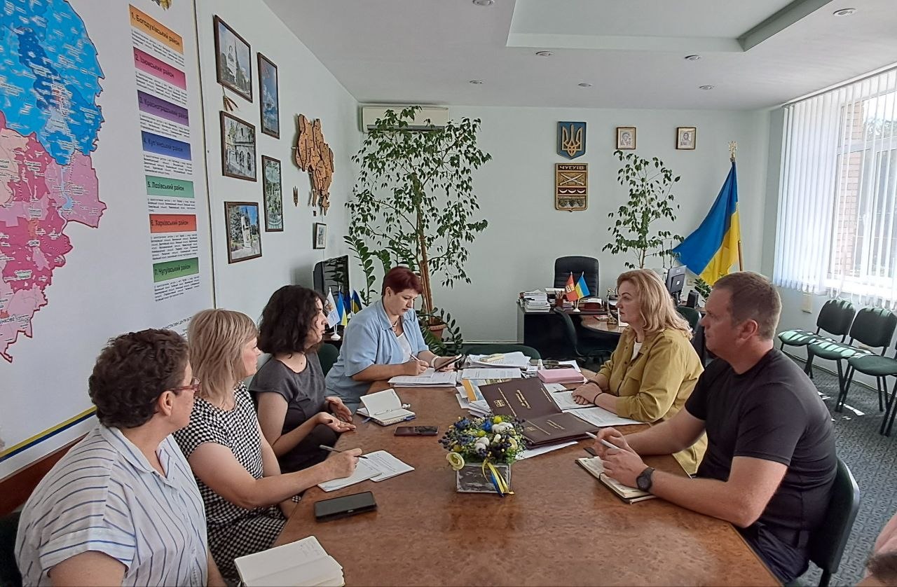 Група людей проводить нараду за конференц-столом, записуючи нотатки, на тлі стіни з адміністративною картою та інформаційним стендом із назвами районів.