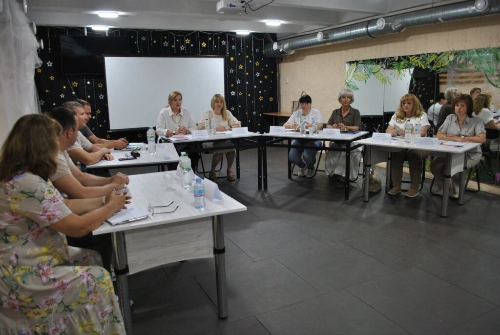 Кілька людей сидять за столами у приміщенні з білою дошкою та проектором на тлі, готуючись до заходу із використанням паперів та пляшок з водою.