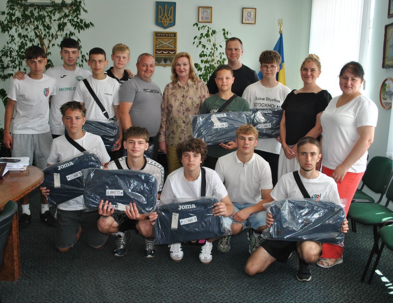 Кілька молодих чоловіків і дорослих стоять і сидять у приміщенні, позуючи для групового фото і тримаючи в руках спортивне приладдя у поліетиленових чохлах із логотипом Joma.