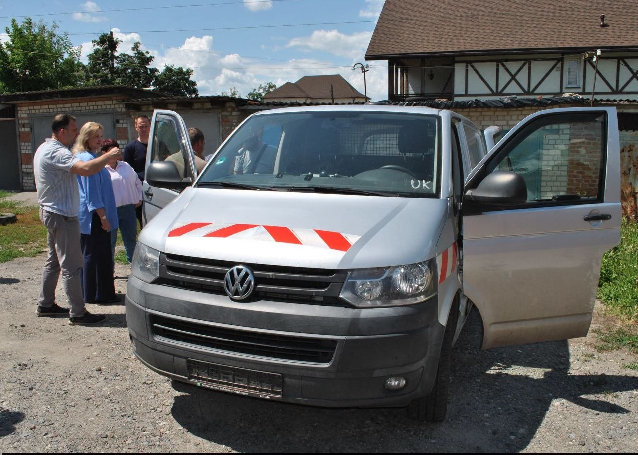 Сріблястий мікроавтобус Volkswagen з відкритими дверима та червоними смугами на капоті припаркований біля будинку, поруч група людей.