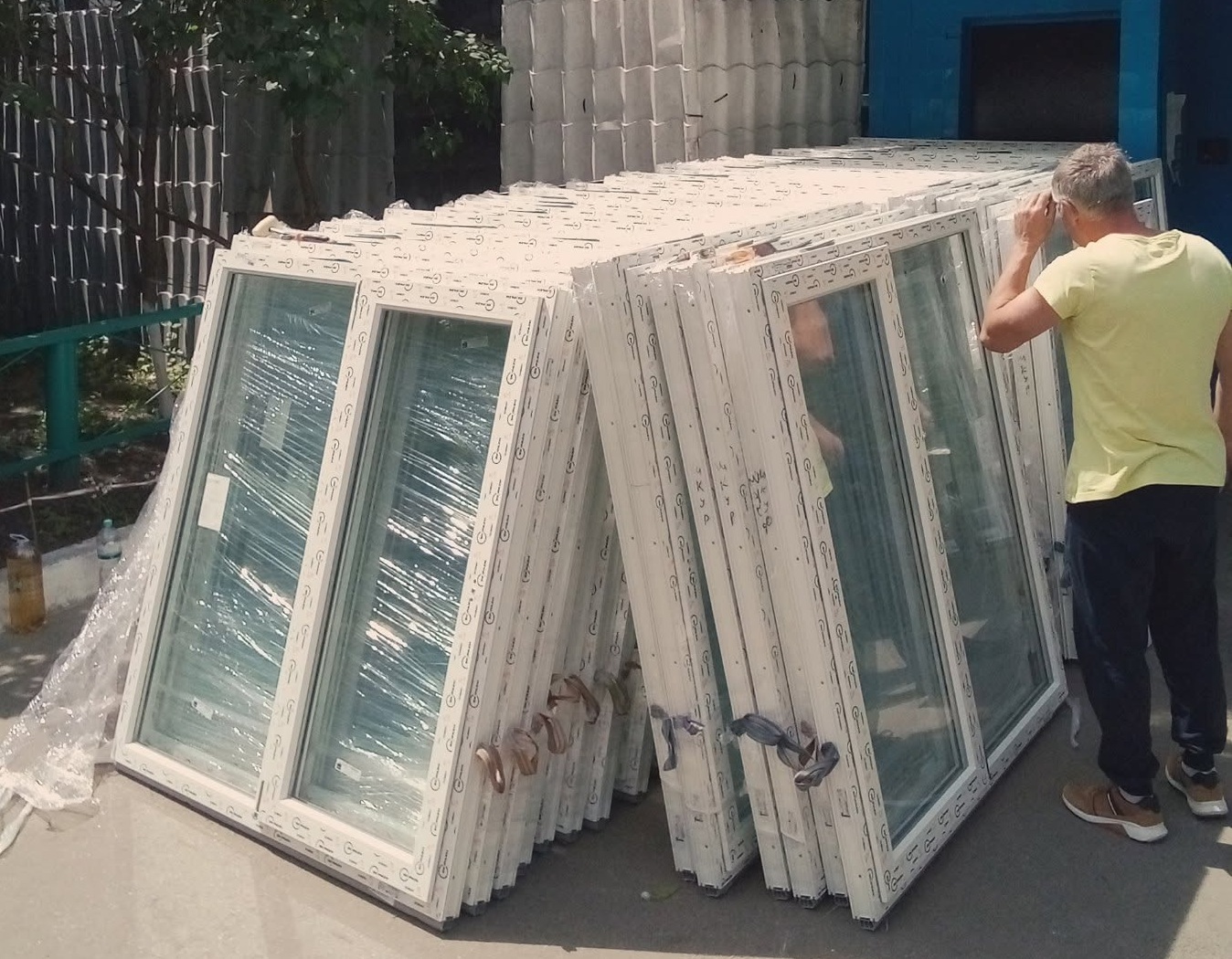 Чоловік у жовтій футболці стоїть поруч із великим стосом нових білих вікон, обгорнутих у захисну плівку.