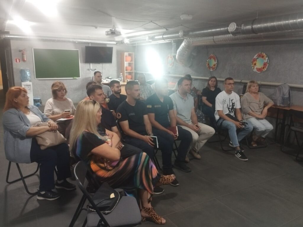 Група людей сидить на стільцях у приміщенні з дошкою та телевізором, ймовірно, відвідуючи лекцію або сем