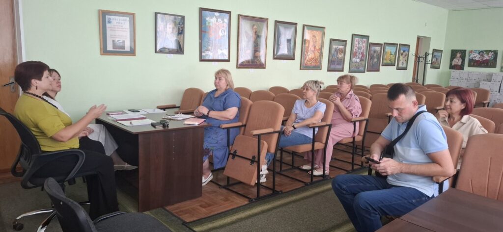 Alternative:* Група людей сидить у світлій кімнаті з іконами на стінах, де жінка в жовтій блузці активно жестикулює під час розмови за столом. (A group of people sits in a light room with icons on the walls, where a woman in a yellow blouse is actively gesturing during a conversation at a table.)