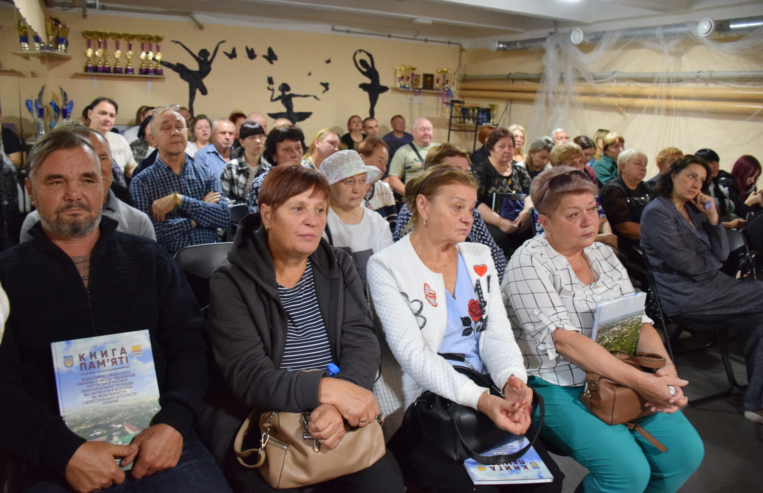 Група людей сидить у залі з трофеями на стіні, а чоловік на передньому плані тримає книгу з назвою 