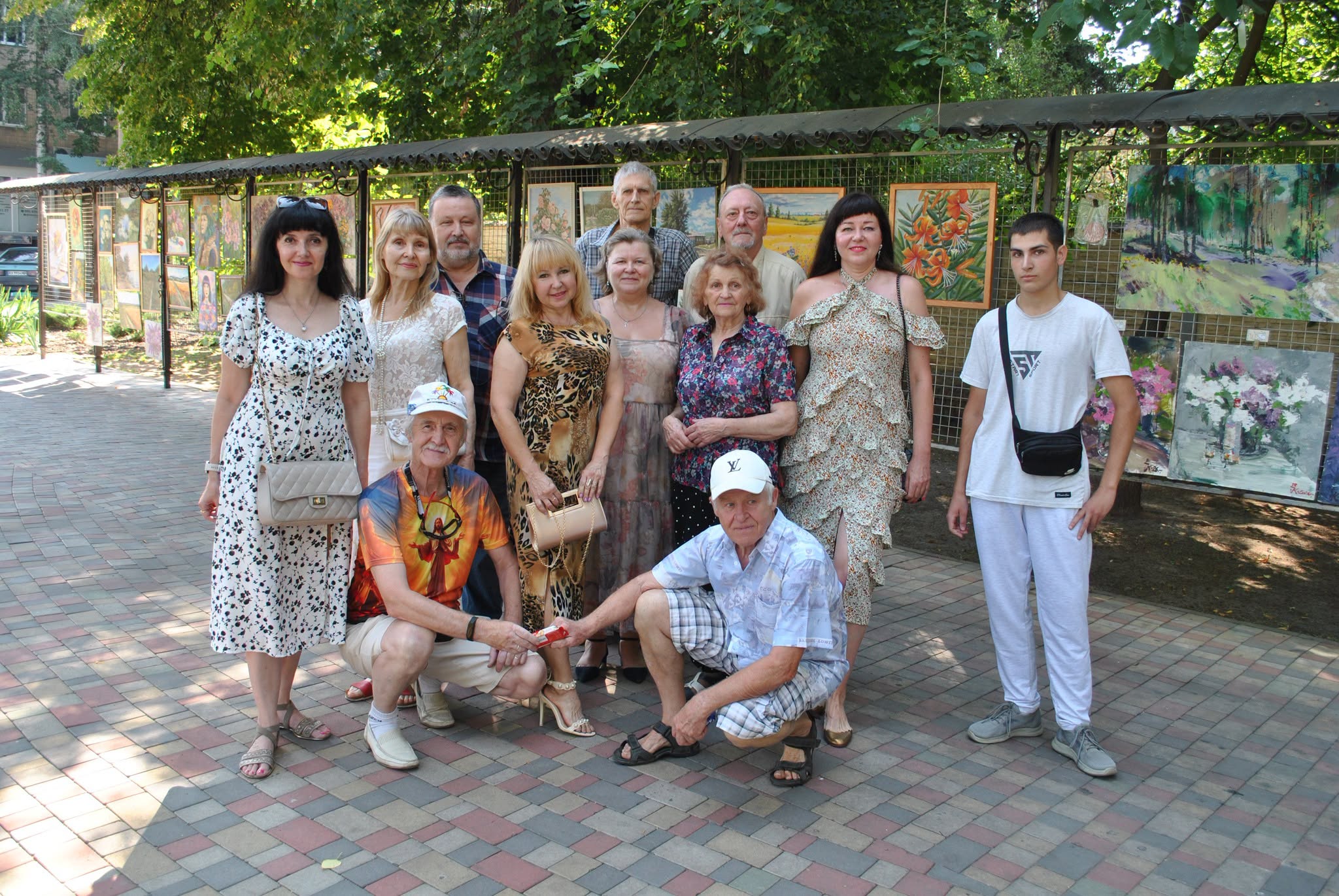 Група людей позує для спільної фотографії на фоні виставки картин під відкритим небом.