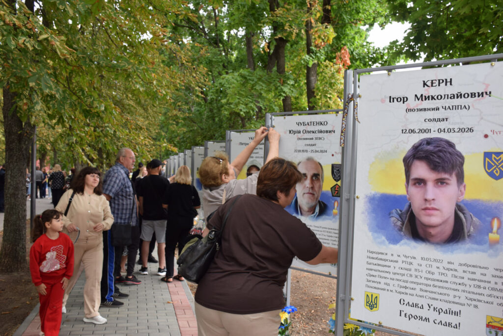 Люди вшановують пам'ять загиблих воїнів біля меморіальної стіни, де жінка торкається портрета солдата Ігоря Керна.