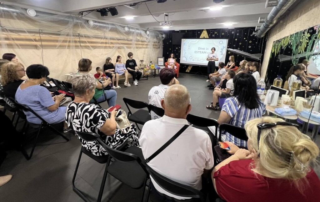 Учасники заходу сидять у залі та дивляться на презентацію, де на екрані відображено напис 'Dive in ESTEAM'.