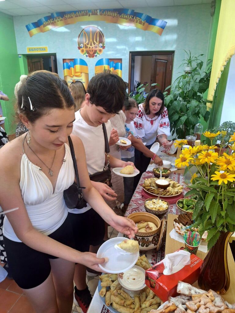 Група людей обслуговує себе їжею з буфету в приміщенні, прикрашеному українськими прапорами та гаслом