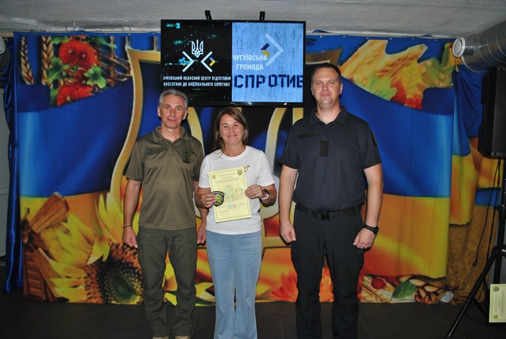 Жінка тримає сертифікат та нагрудний знак, стоячи між двома чоловіками перед екраном із написом