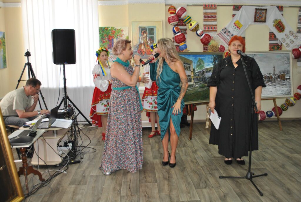 Жінки виступають з мікрофонами на культурному заході, супроводжувані звукорежисером та традиційними українськими декораціями.