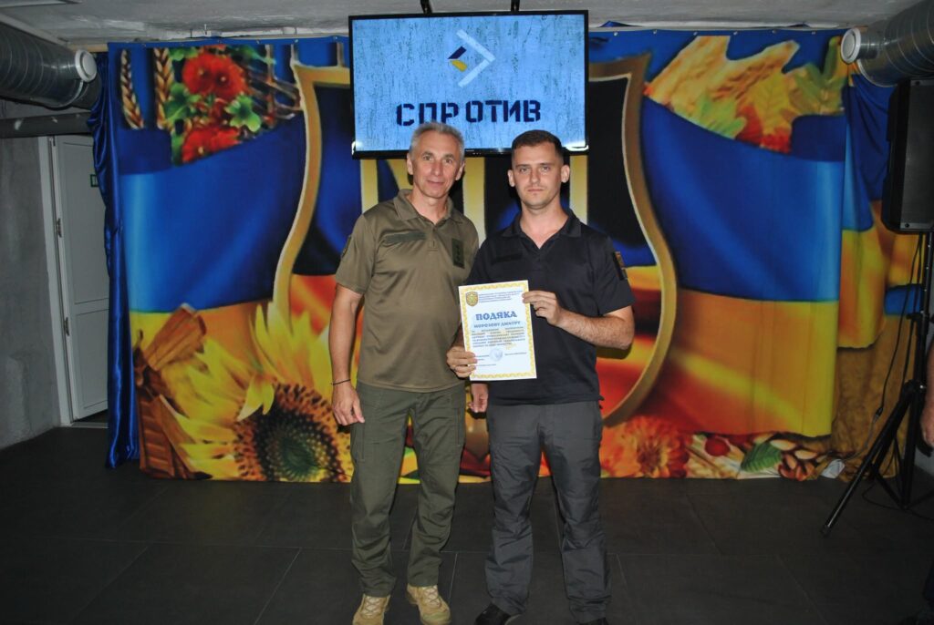 Два чоловіки стоять на фоні українських прапорів та екрана з написом