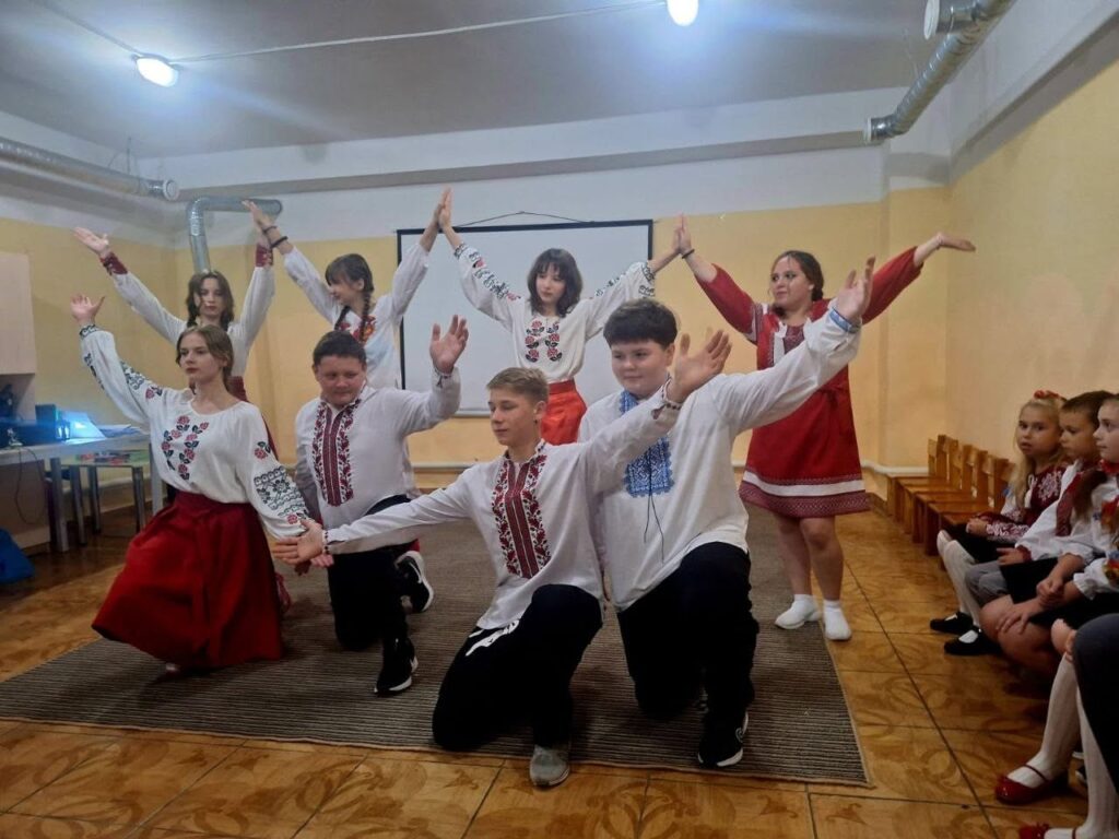 Група дітей та підлітків у традиційних вишиванках виконує народний танець у приміщенні.