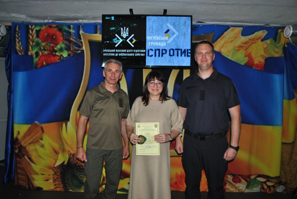 Жінка тримає сертифікат та нагрудний знак, стоячи між двома чоловіками перед екраном з написом «Арківський обласний центр підготовки населення до національного спротиву».