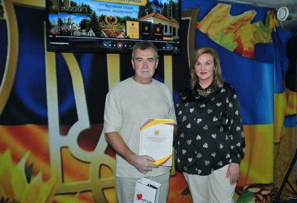 Чоловік тримає грамоту та подарункову сумку з логотипом Чугуєва, стоячи поруч із жінкою на церемонії нагородження.