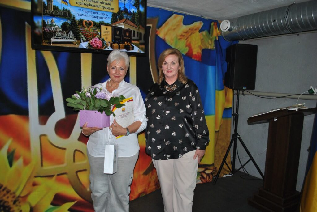 Дві жінки стоять на фоні зображення карти України, при цьому жінка зліва тримає букет квітів та грамоту, а на екрані вище видно напис про вручення подяк та подарунків у Ч