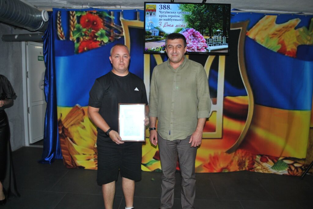 Два чоловіки стоять перед банером із прапором України, при цьому чоловік зліва тримає рамку з документом, а на екрані позаду них відображено напис «388. Чугуївська громада крізь героїзм — до майбутнього! З Днем міста!».