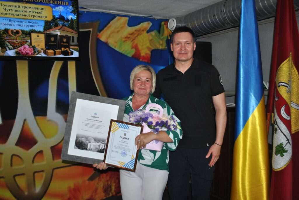 Жінка тримає рамку з подякою та букет квітів, стоячи поруч із чоловіком у чорній формі на тлі українського прапора та екрана з написом про вручення нагород.