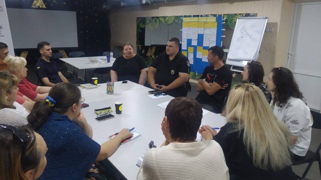 Група людей сидить навколо білого столу під час ділової зустрічі, обговорюючи питання та роблячи нотатки.