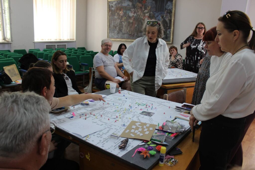 Група людей обговорює велику схему на столі, розміщуючи на ній цвяхи та стикери.