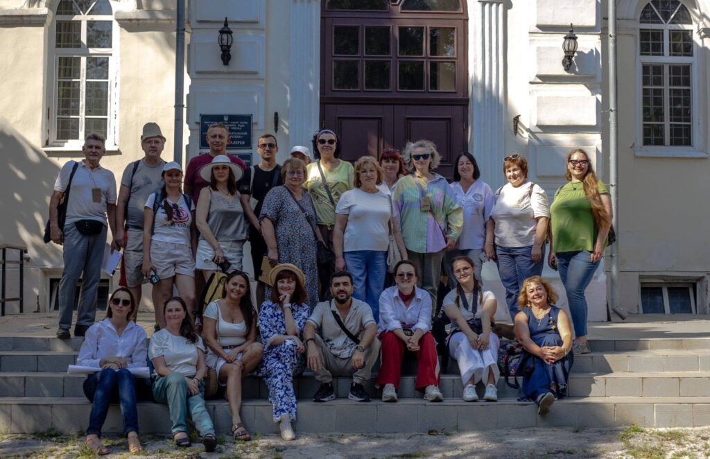 Група людей стоїть і сидить на сходах перед будівлею з великими дерев'яними дверима.