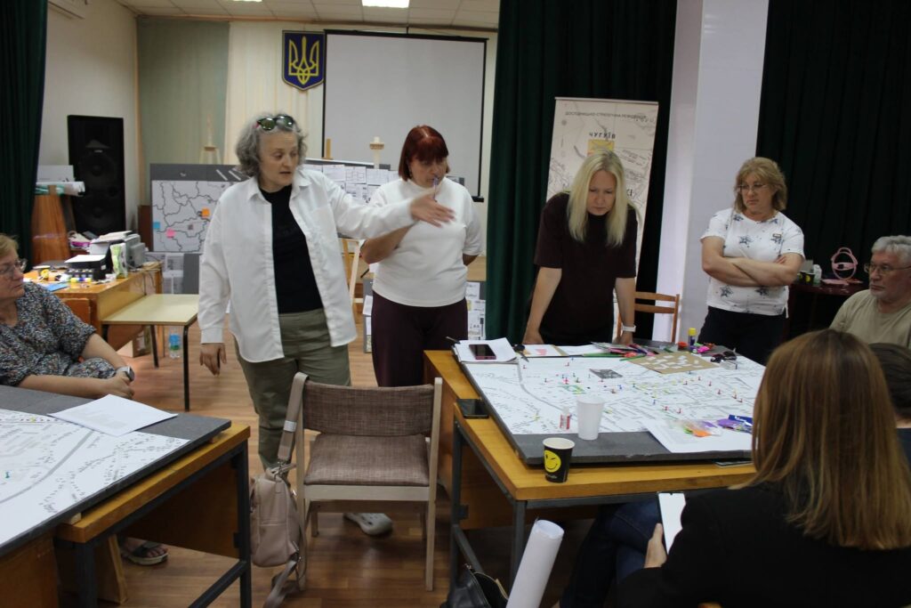 Учасники зустрічі обговорюють великі плани та карти, розкладені на столах.