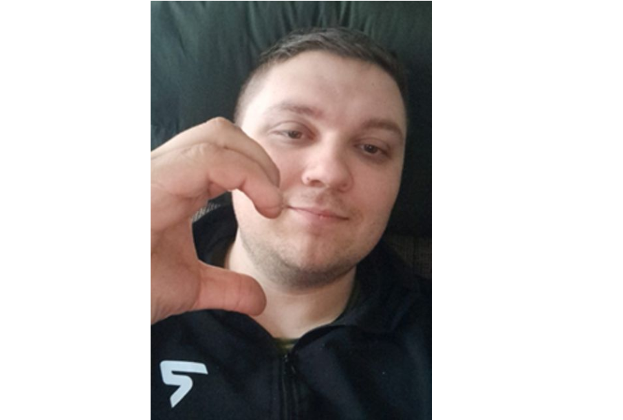 Молодий чоловік у чорній куртці з білим логотипом робить жест рукою біля обличчя.