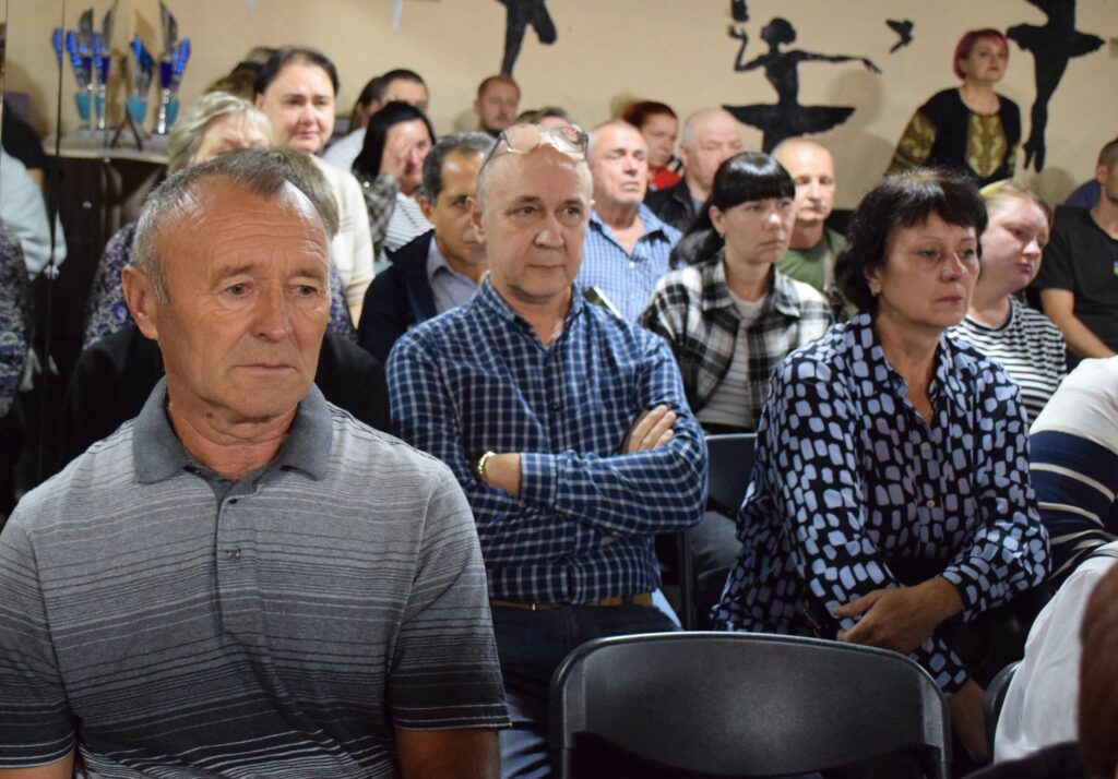 Чоловік у сірій смугастій футболці сидить у залі, дивлячись вперед, позаду нього інші учасники заходу.