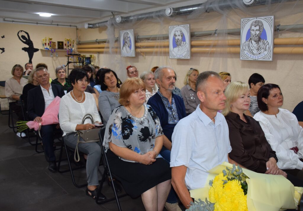 Учасники заходу сидять у залі, прикрашеному портретами, зокрема Геродота, та трофеями.