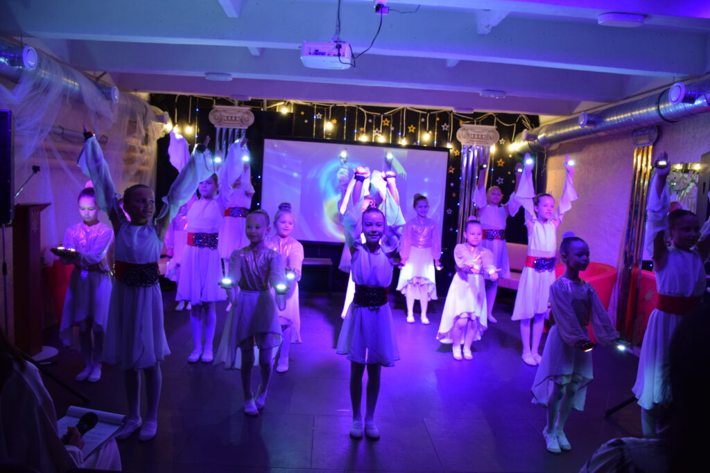 Група дівчаток у білих сукнях виконує виступ на сцені, тримаючи світлові палички та прозорі тканини.