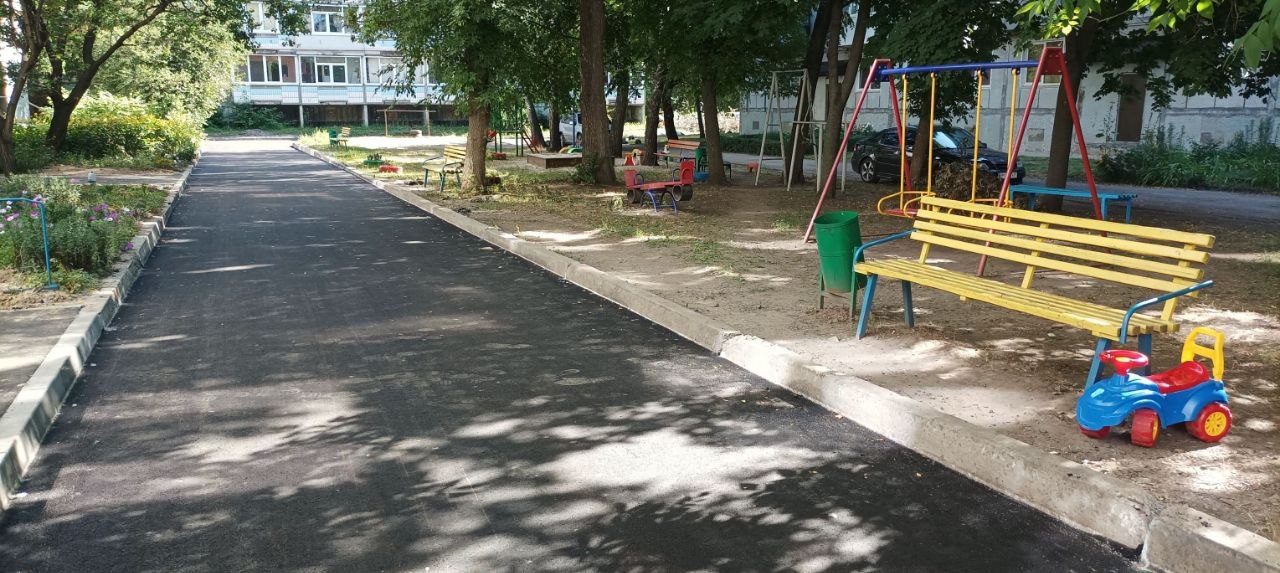 Асфальтована доріжка в парку, поруч з якою розташовані жовта лавка, зелений сміттєвий бак та іграшкова машинка під тінню дерев.