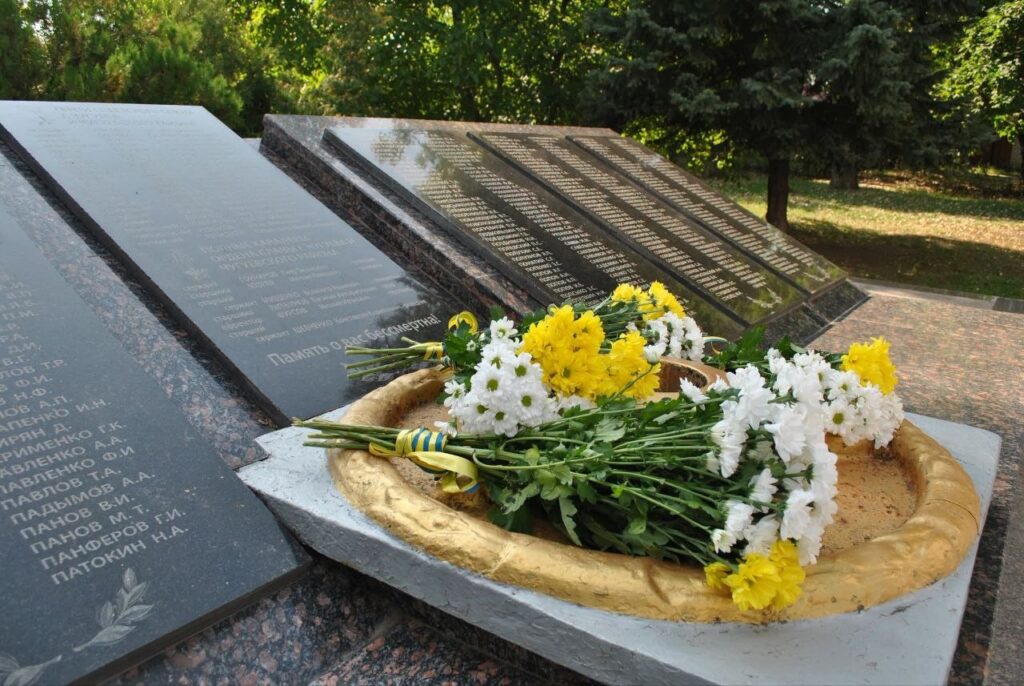 Чорні гранітні плити меморіалу з написом «Пам'ять» та списками імен, біля яких у золотій чаші вічного вогню лежать білі та жовті хризантеми з синьо-жовтою стрічкою.