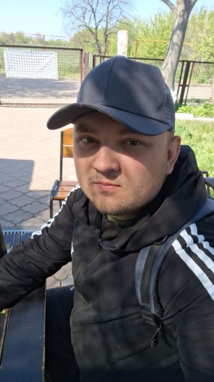 Чоловік у темній кепці та чорному худі з білими смугами сидить на лавці на вулиці.