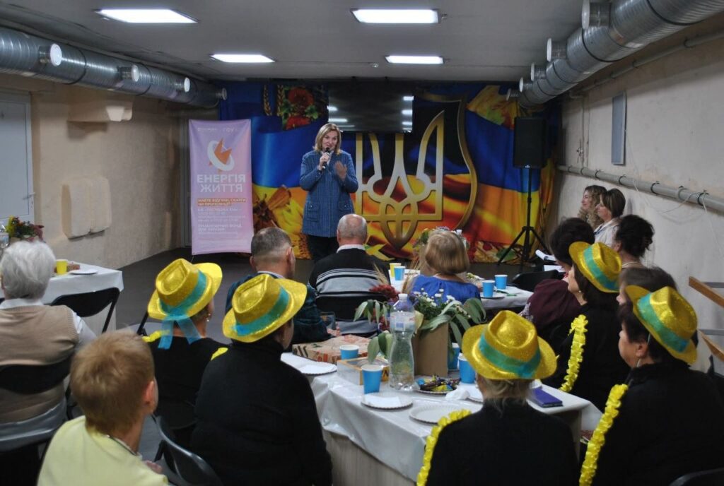 Жінка виступає з мікрофоном перед аудиторією в залі з українським прапором та банером «Енергія життя».