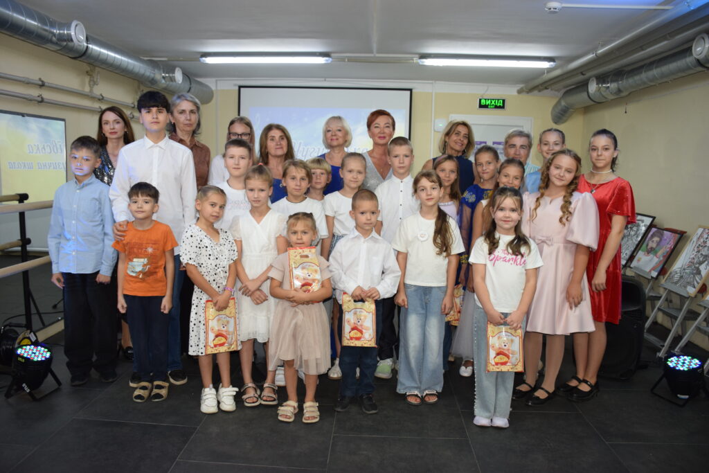 Група дітей та дорослих позує для спільного фото, деякі діти тримають подарунки, на тлі видно вивіску 