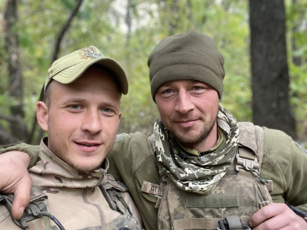 Два військовослужбовці в камуфляжі обіймають одне одного на тлі лісу.