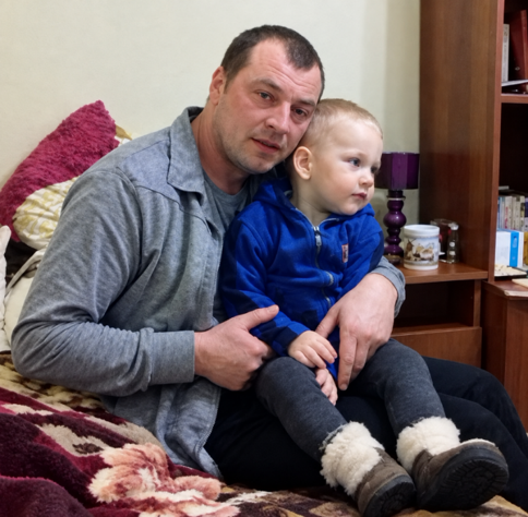 Чоловік у сірій сорочці тримає на колінах малюка в синій куртці та вовняних чобітках.