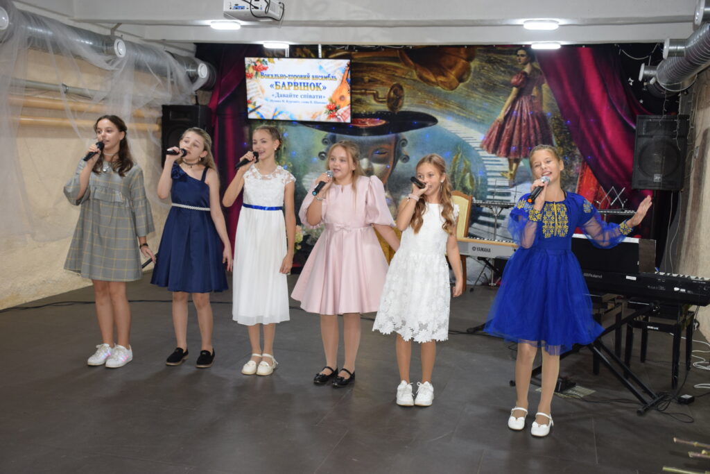 Шестеро дівчат у святкових сукнях співають у мікрофони на сцені під час концерту вокально-танцювального ансамблю «Барвінок».