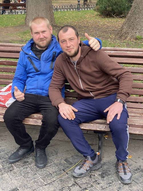 Двоє чоловіків сидять на парковій лавці, один із них робить жест великим пальцем вгору.