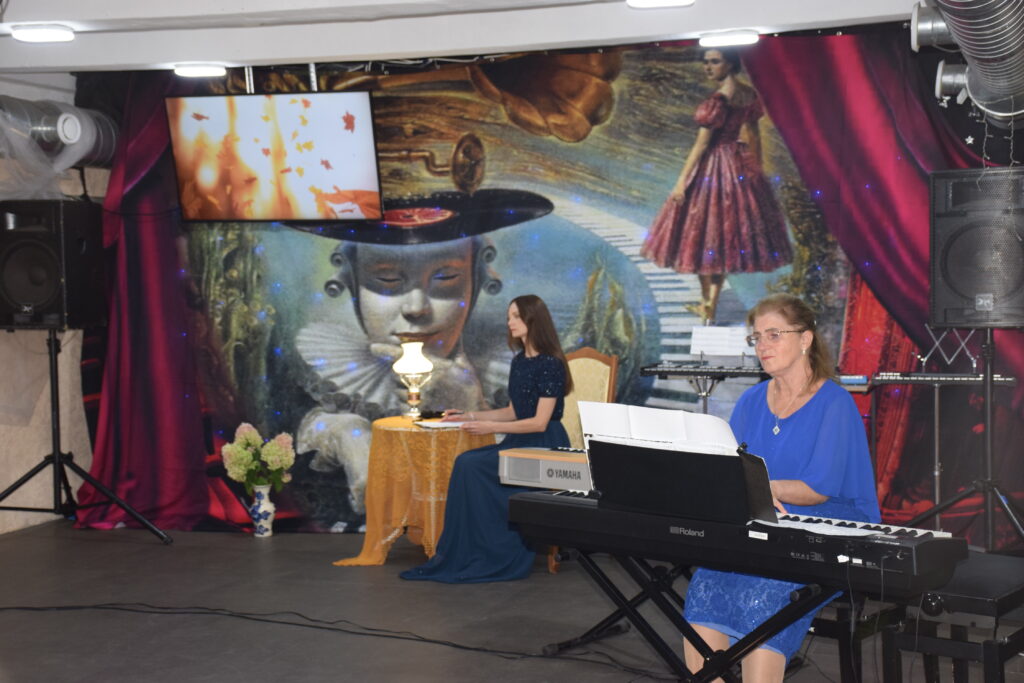 Дві жінки грають на електронних піаніно Yamaha та Roland на сцені з яскравим фоновим екраном.